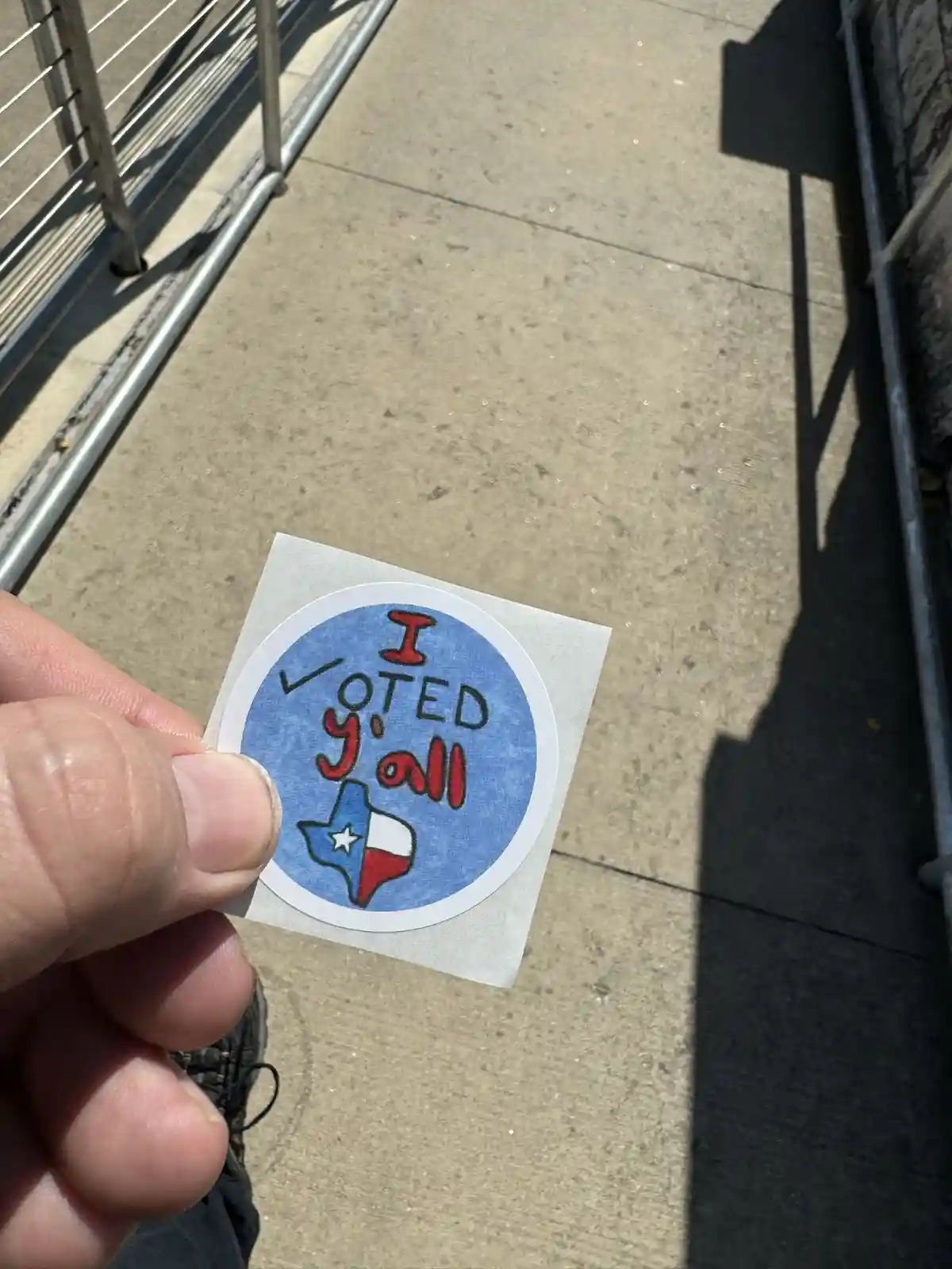 A hand holds a circular sticker with a blue background, featuring the text 'I VOTED y'all' in red and black lettering, with…