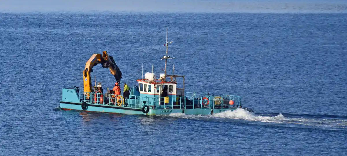 A light blue workboat named 'MEERCAT' sails on calm blue water, carrying a large orange excavator arm and two workers in…