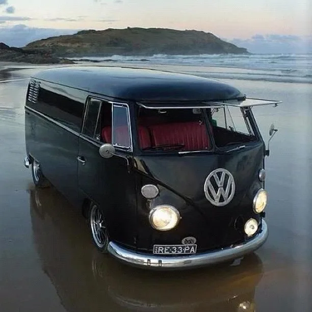 A black vintage Volkswagen Type 2 van is parked on wet sand at a beach during sunset or sunrise, with waves and a distant…