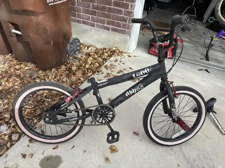 A black Kent BMX bike with pink-rimmed wheels is parked on a concrete driveway next to a brown trash bin and a pile of…