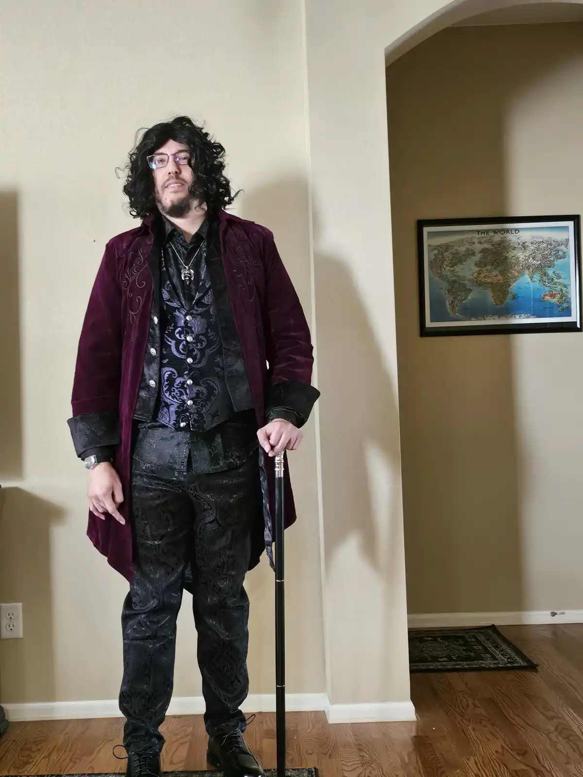 A man dressed in a flamboyant, dark purple velvet coat over a black patterned suit, holding a cane, stands indoors with a…