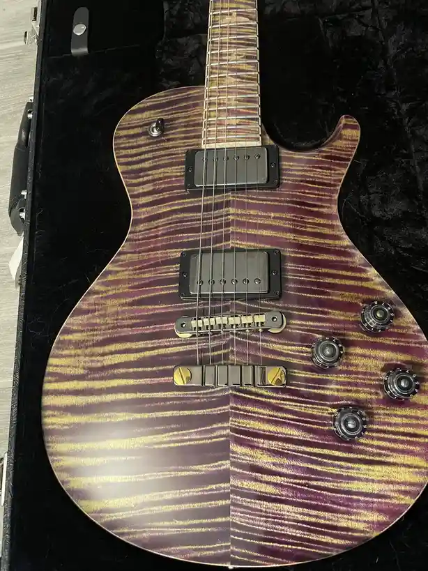 a close-up photo of an electric guitar with a distinctive purple and gold striped finish, resting inside a black…