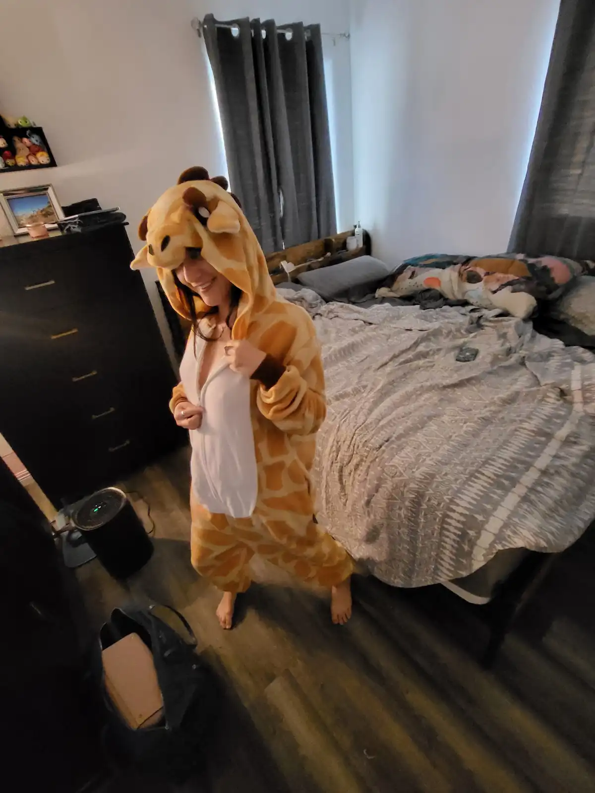 A person in a full-body giraffe onesie stands in a bedroom, smiling and pulling open the front zipper to reveal a white…