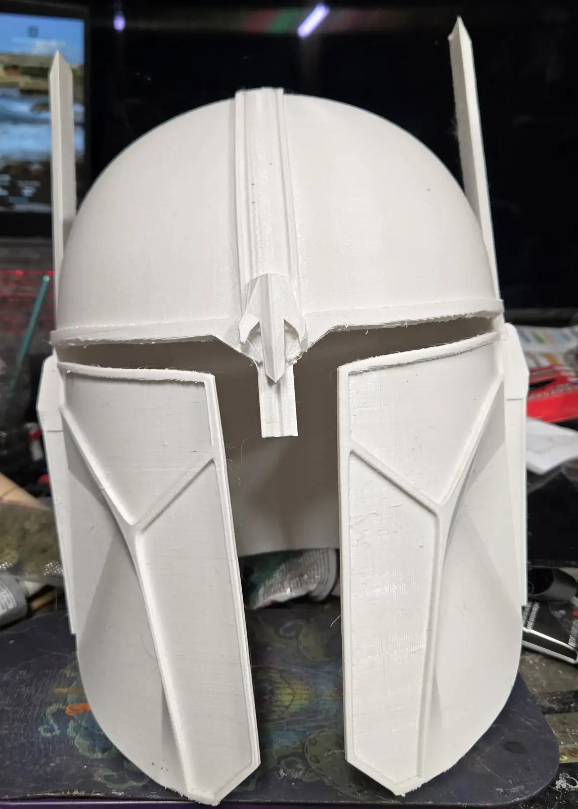 A white, unfinished 3D-printed Mandalorian helmet sits on a cluttered work surface, showing visible layer lines and rough…