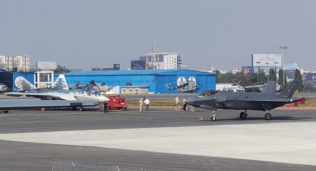 Two fighter jets, an F-35 on the right and a Su-57 on the left, are parked on an airfield tarmac with people and other…