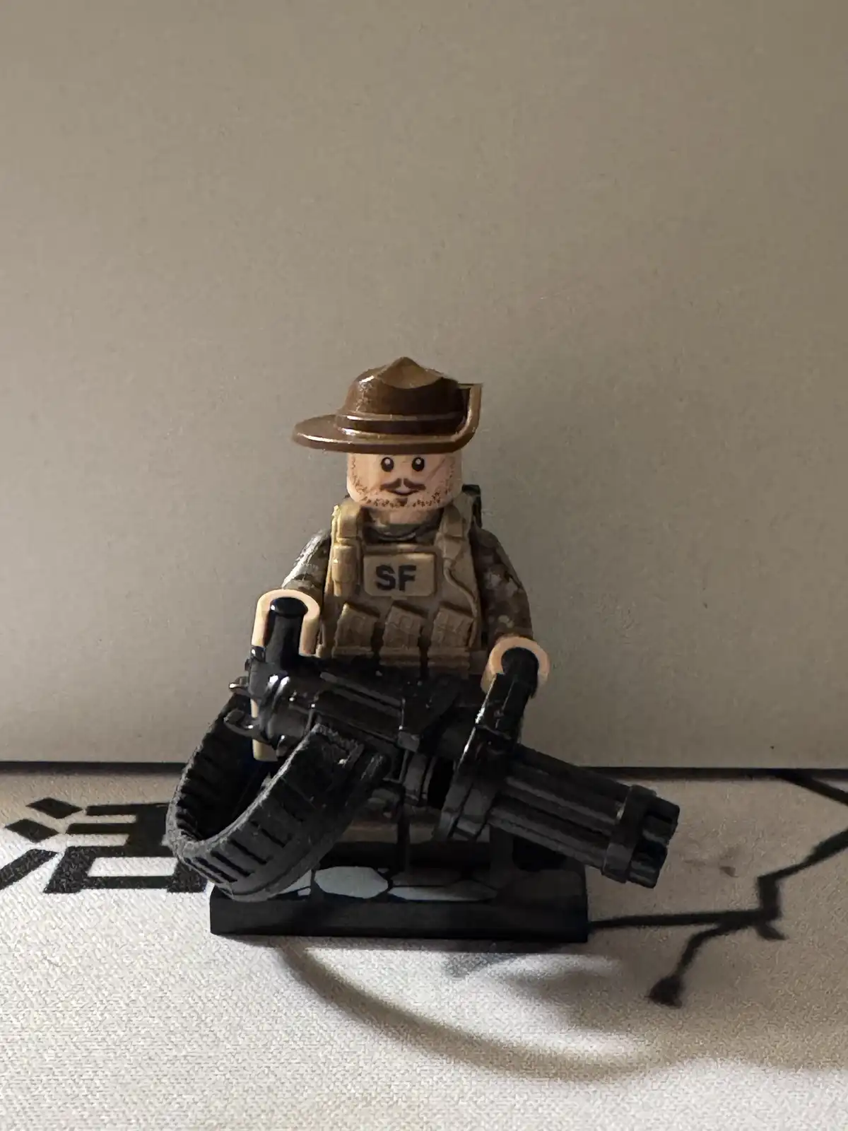 A custom-modified LEGO minifigure dressed as a soldier with a brown hat, camouflage vest marked 'SF', and holding a large…