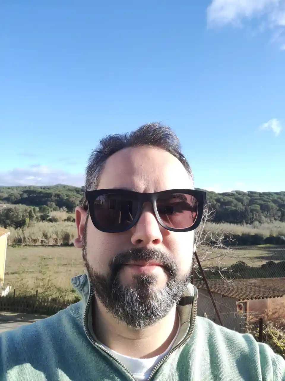 A man with a beard and sunglasses takes a selfie outdoors under a clear blue sky, with hills and vegetation in the…