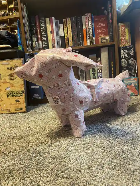 A handmade dog-shaped pinata, crafted from pink wrapping paper decorated with candy cane and bow patterns, stands on a…