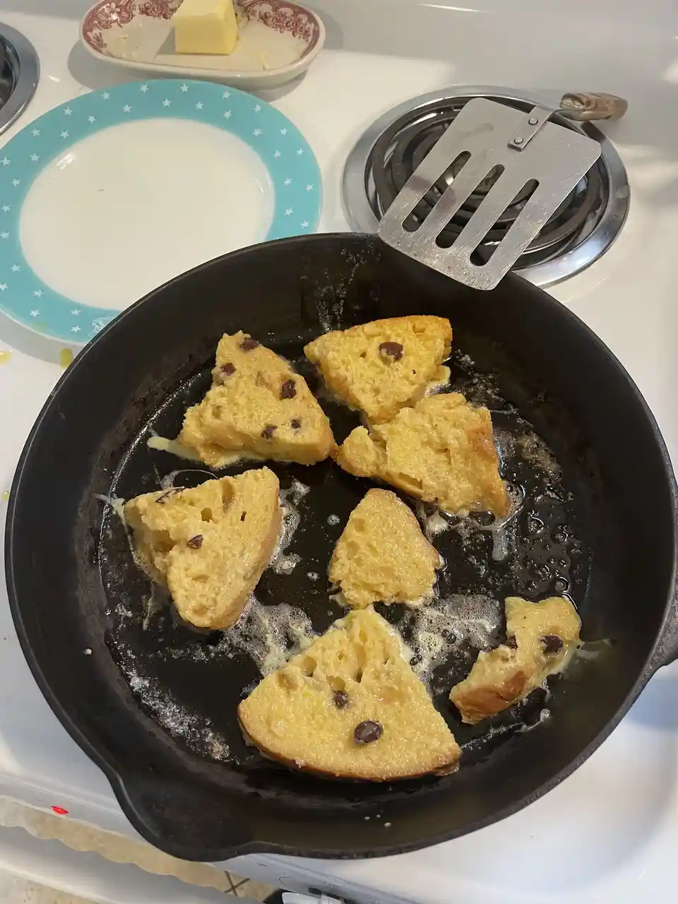 A black skillet on a stovetop contains several golden-brown, triangular pieces of French toast sizzling in melted butter. A…