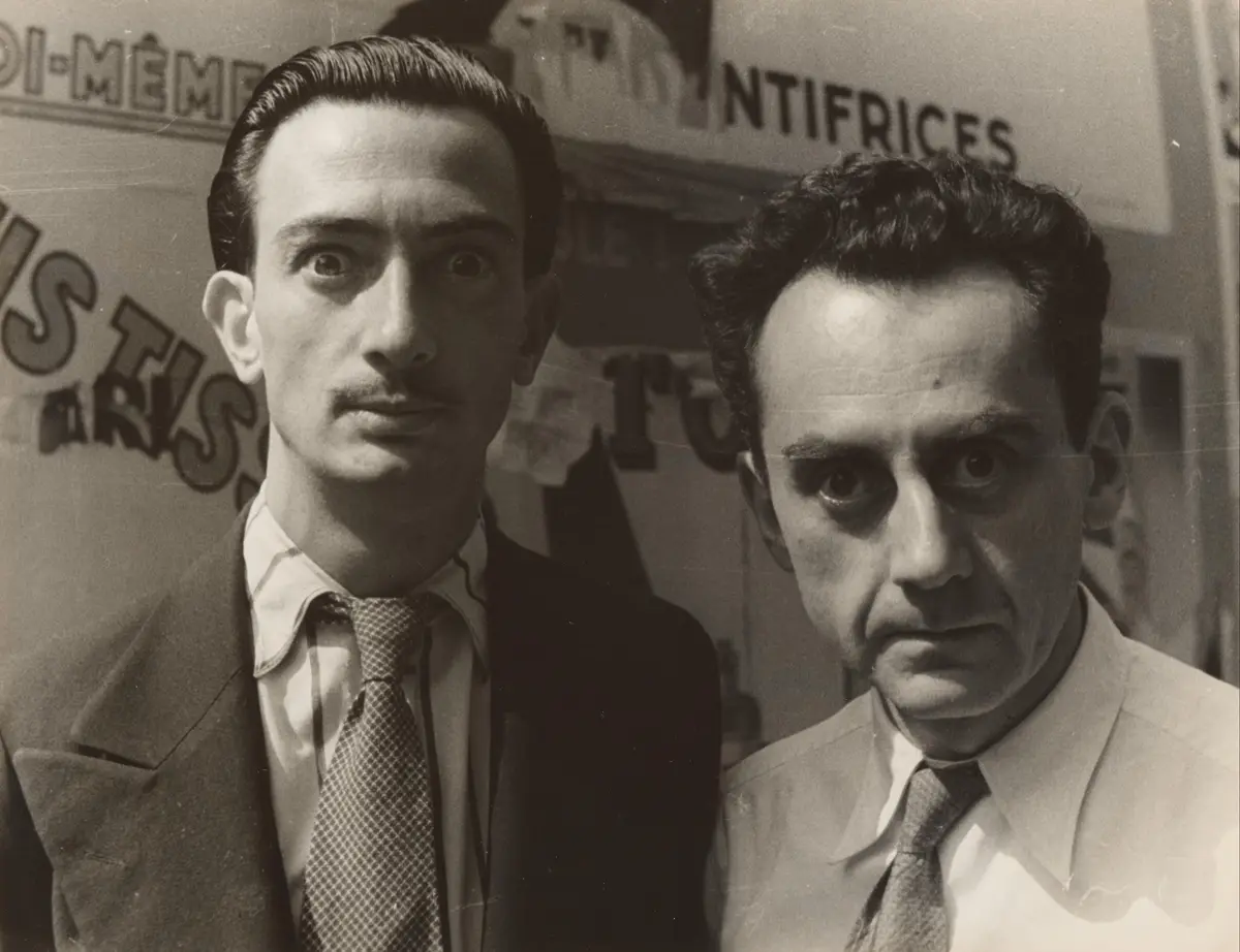 A black-and-white photo shows two men in suits looking directly at the camera with intense, unamused expressions. The man on…