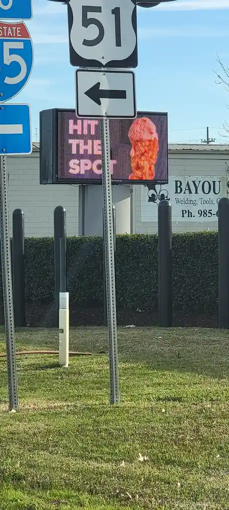A roadside digital sign displays the text 'HIT THE SPOT' in pink letters next to a pixelated orange face, mounted below a…