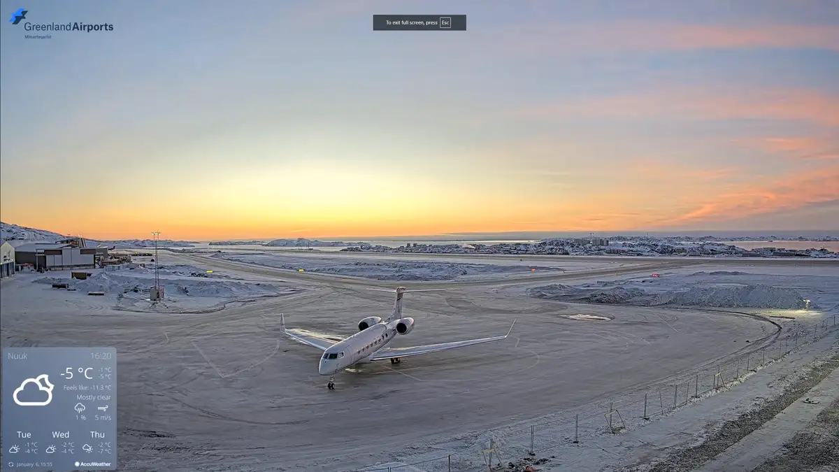 A wide shot of an airport tarmac in Nuuk, Greenland, covered in snow, with a small white airplane parked on the runway…