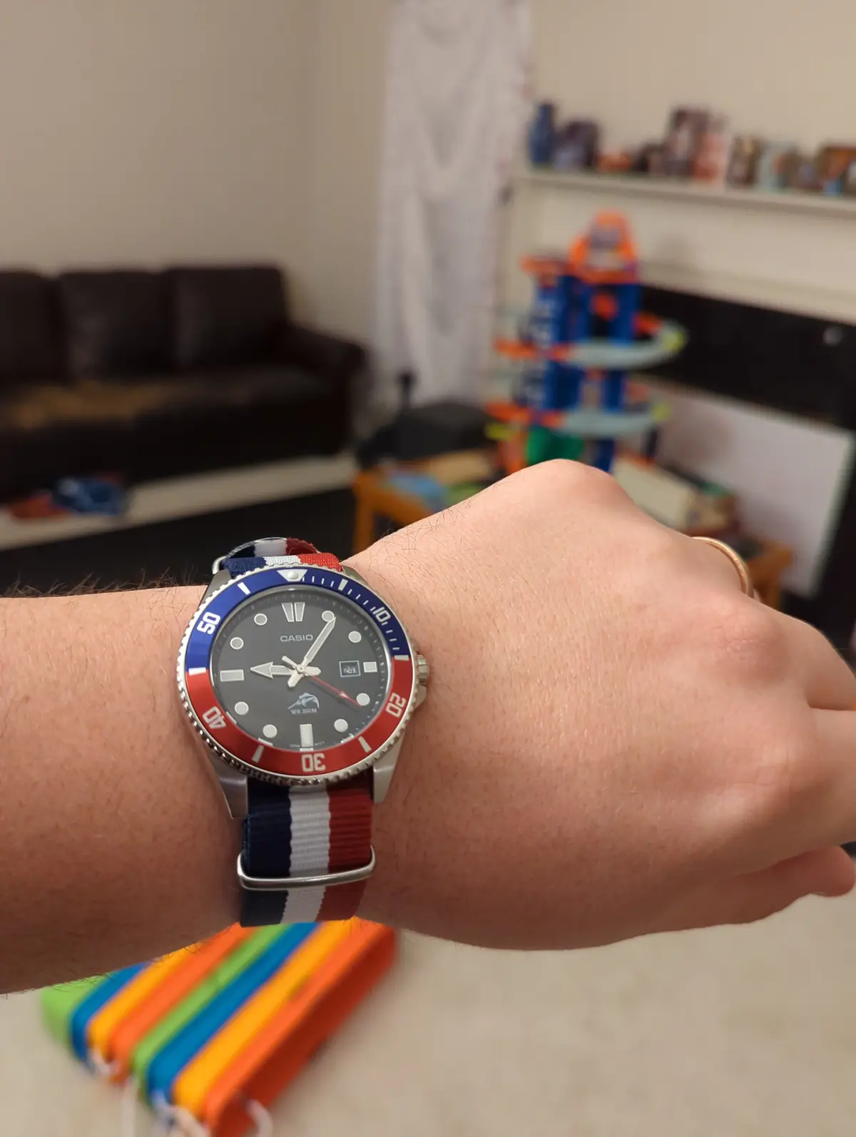 A close-up photo of a person's wrist wearing a Casio watch with a red, white, and blue striped NATO strap and a matching red…