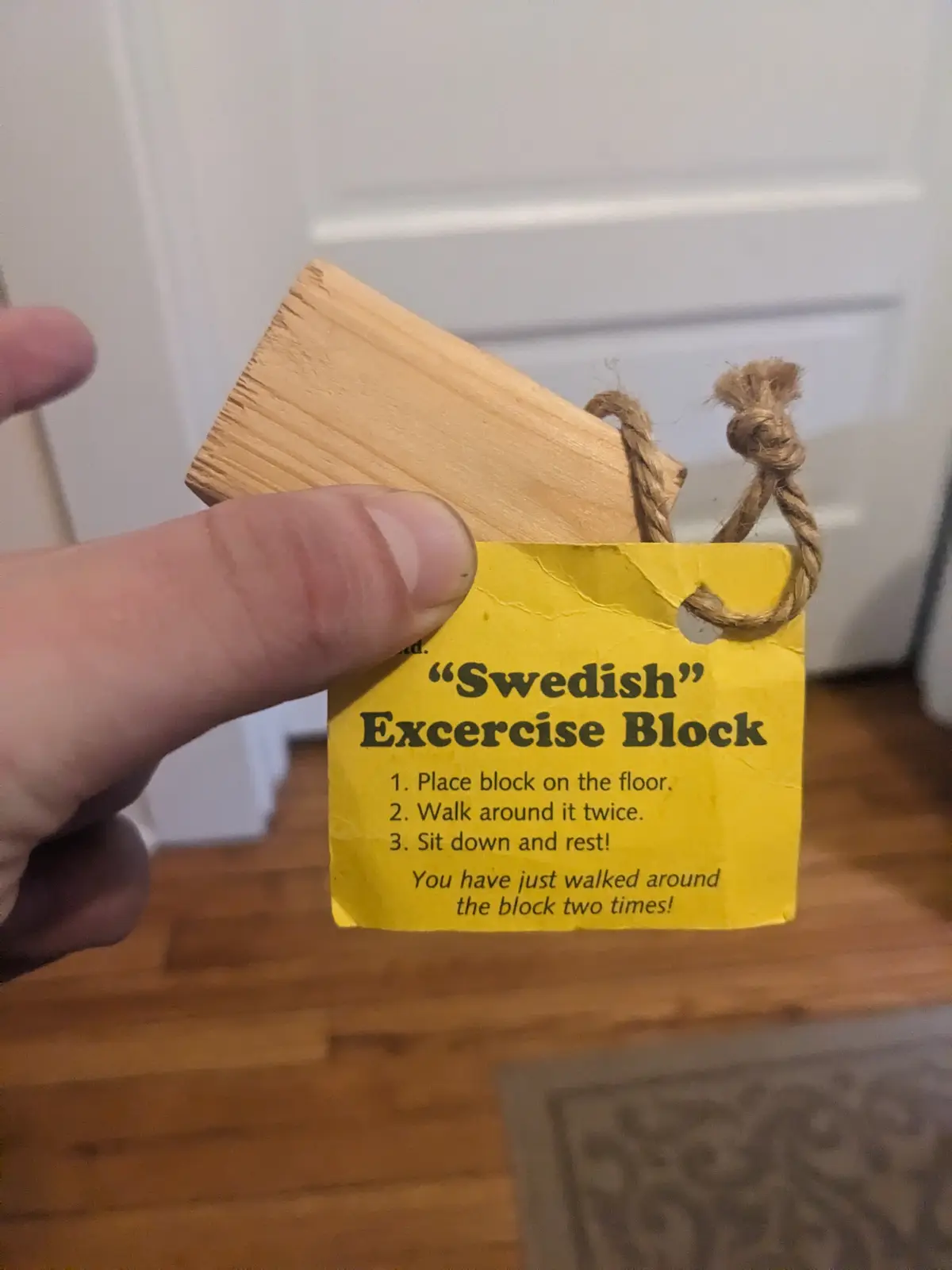 A hand holds a small wooden block with a yellow tag attached by twine. The tag reads "Swedish" Exercise Block and lists…
