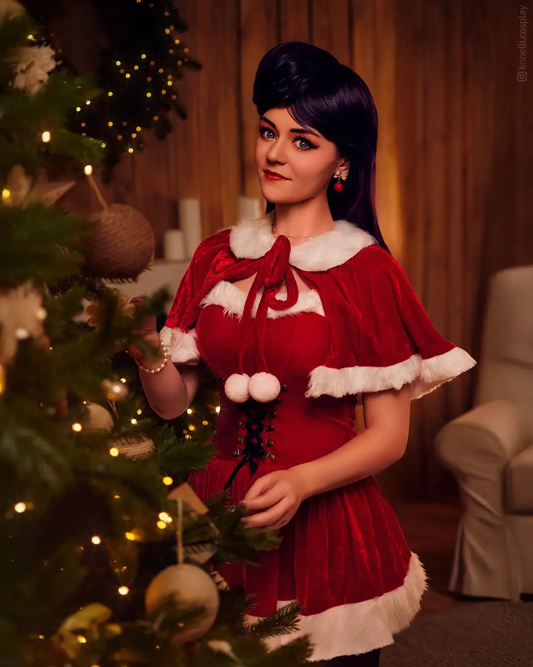 A woman with dark hair and blue eyes wears a red velvet dress with white fur trim and a matching cape, standing beside a…