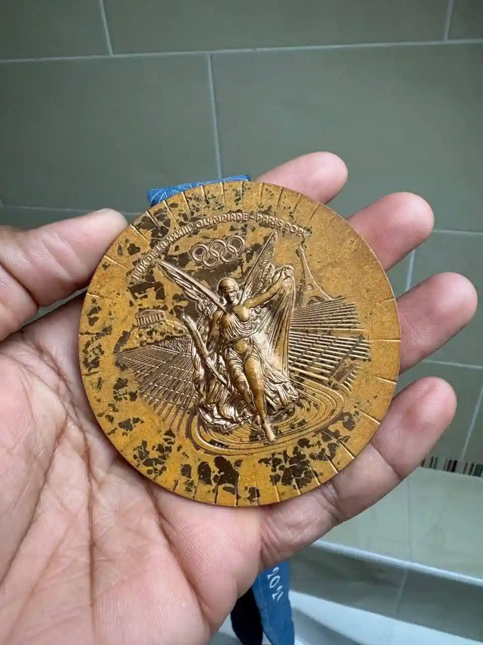 a hand holds a gold-colored Olympic medal from the 2024 Paris Games, which is visibly covered in black, soot-like stains…