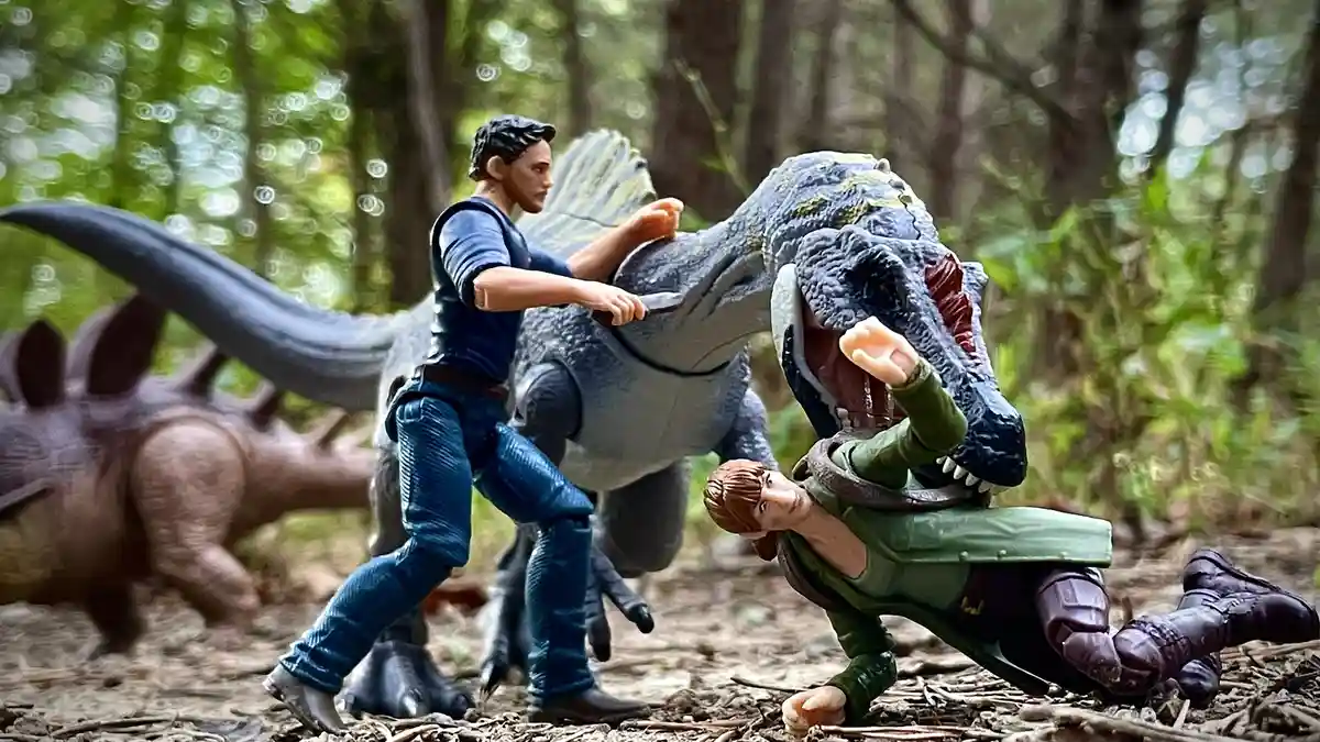 A photo of action figures staged in a forest: one figure in blue is holding onto a grey dinosaur's head while another figure…