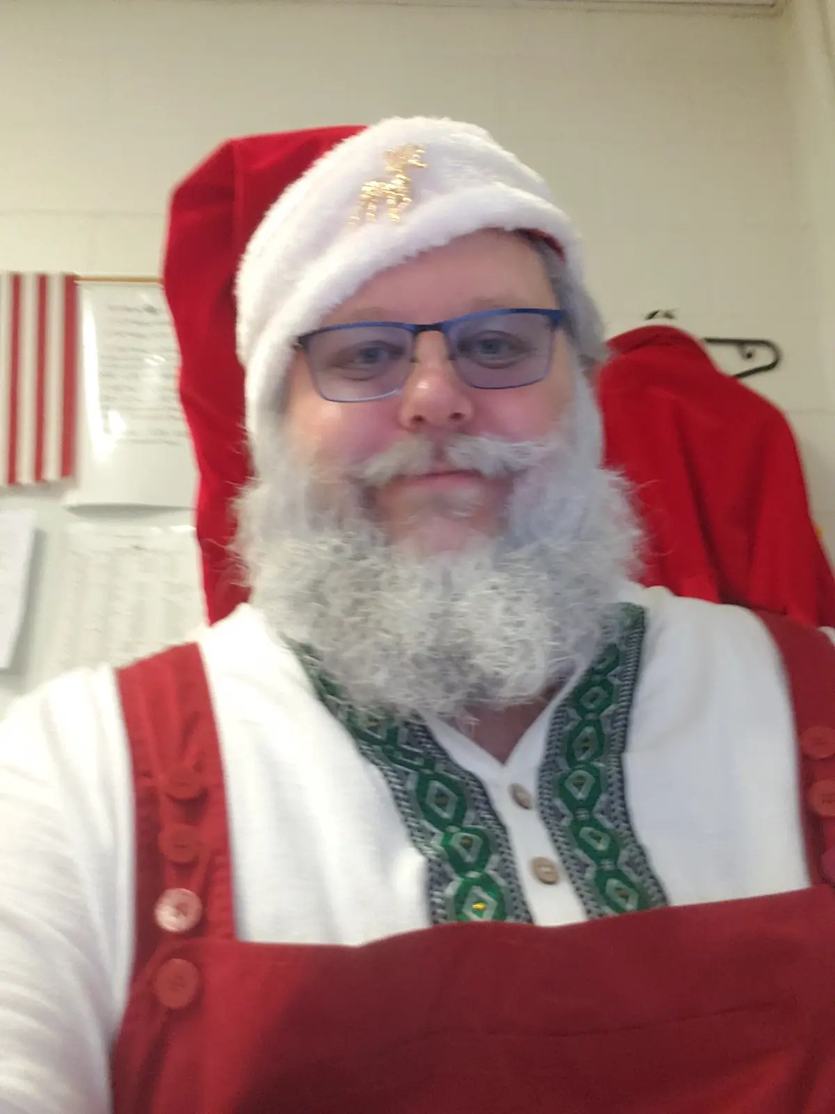 A man wearing a red and white Santa hat with a gold reindeer pin, glasses, and a full white beard, dressed in a red vest…