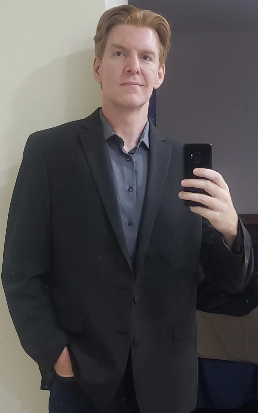 A man with reddish hair takes a mirror selfie wearing a black suit jacket over a grey button-up shirt, holding a black…