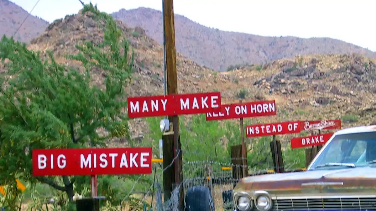 A vintage photo shows a rusty car parked near a wooden post with several red signs. The signs read 'MANY MAKE RELY ON HORN'…