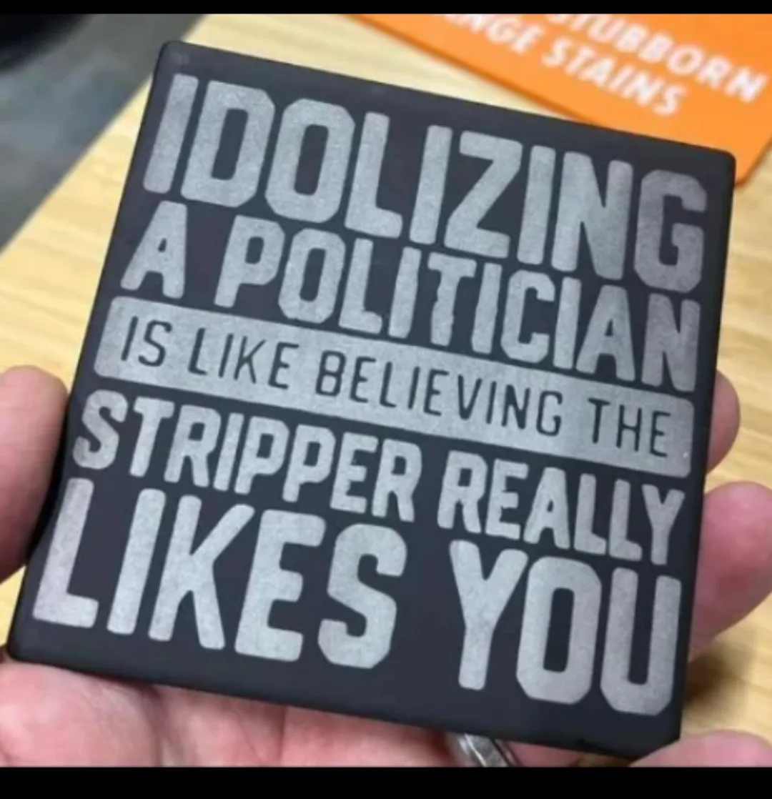 A hand holds a black square coaster with large white text that reads 'IDOLIZING A POLITICIAN IS LIKE BELIEVING THE STRIPPER…