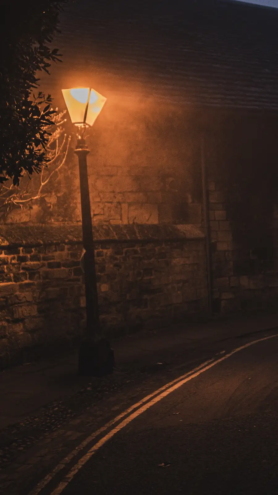 A dimly lit street scene at night, featuring a single vintage-style streetlamp casting a warm orange glow on a cobblestone…