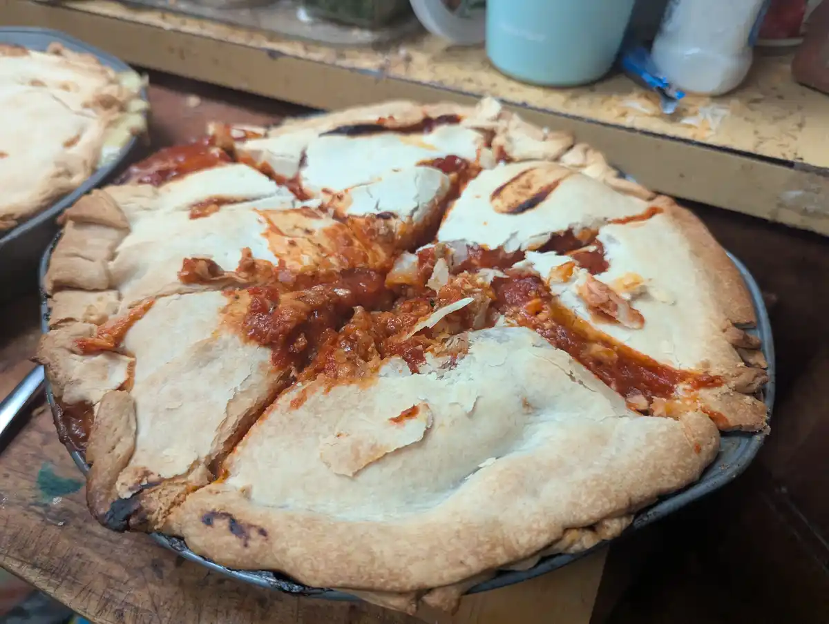 A close-up photo shows a baked dish in a metal pie tin, resembling a deep-dish pizza with a thick crust and tomato sauce…