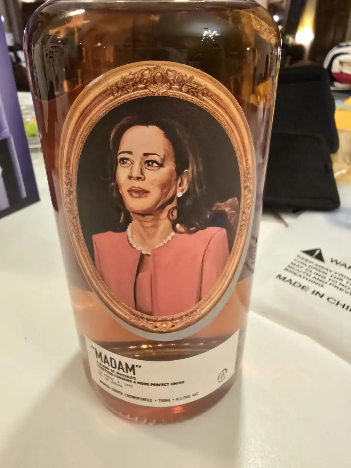 A close-up photo of a whiskey bottle with a portrait of Kamala Harris in an ornate gold frame on its label. The bottle's…