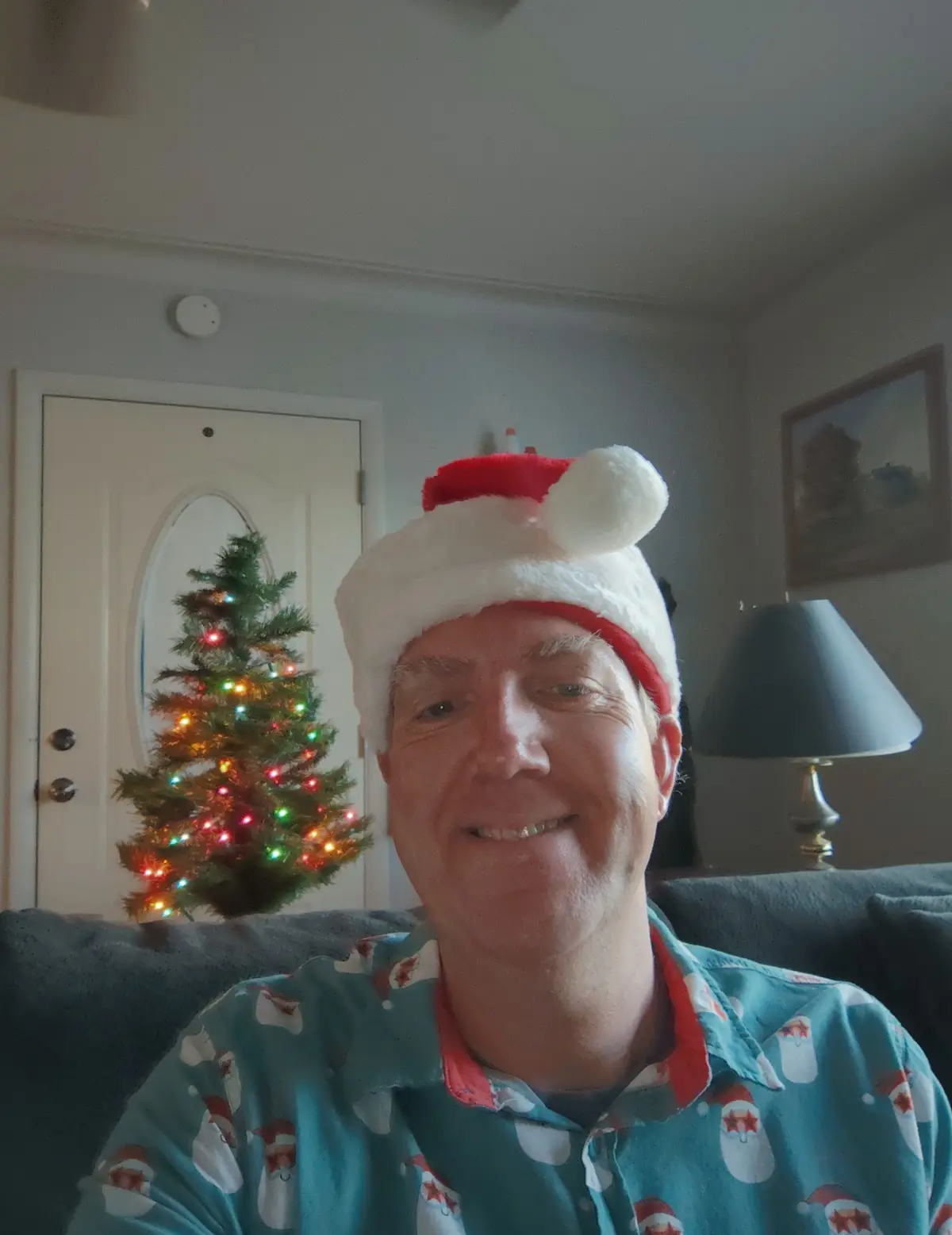 A man wearing a red and white Santa hat and a blue shirt with Santa Claus patterns smiles at the camera while sitting on a…