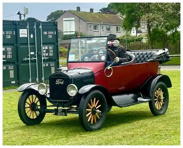 A man in a vintage Ford Model T, wearing a top hat and white gloves, sits in the driver's seat with a large, round, white…