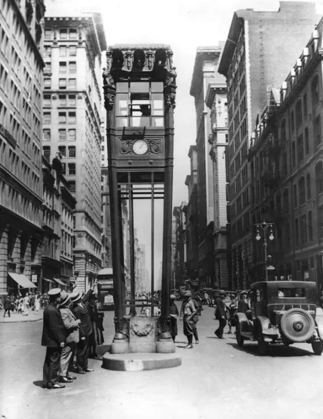 A black-and-white historical photo shows a tall, ornate clock tower standing in the middle of a busy city street lined with…