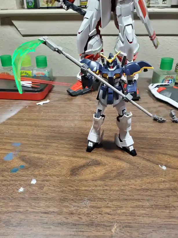 A blue and white plastic model kit robot stands on a wooden table holding a long pole with a green translucent curved blade…