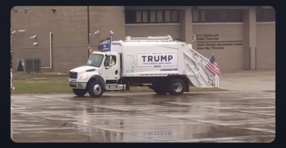 A white garbage truck with 'TRUMP 2024' written on its side is parked on a wet tarmac in front of a building labeled 'U.S…