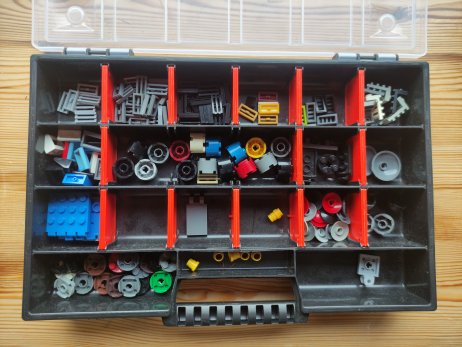 An open black plastic storage box with red dividers, filled with various small Lego pieces like wheels, axles, and plates…