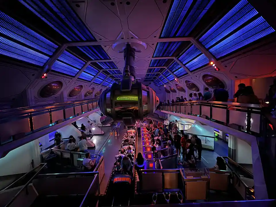 A wide-angle photo shows the interior of a futuristic, sci-fi themed ride queue with blue and purple lighting, featuring a…