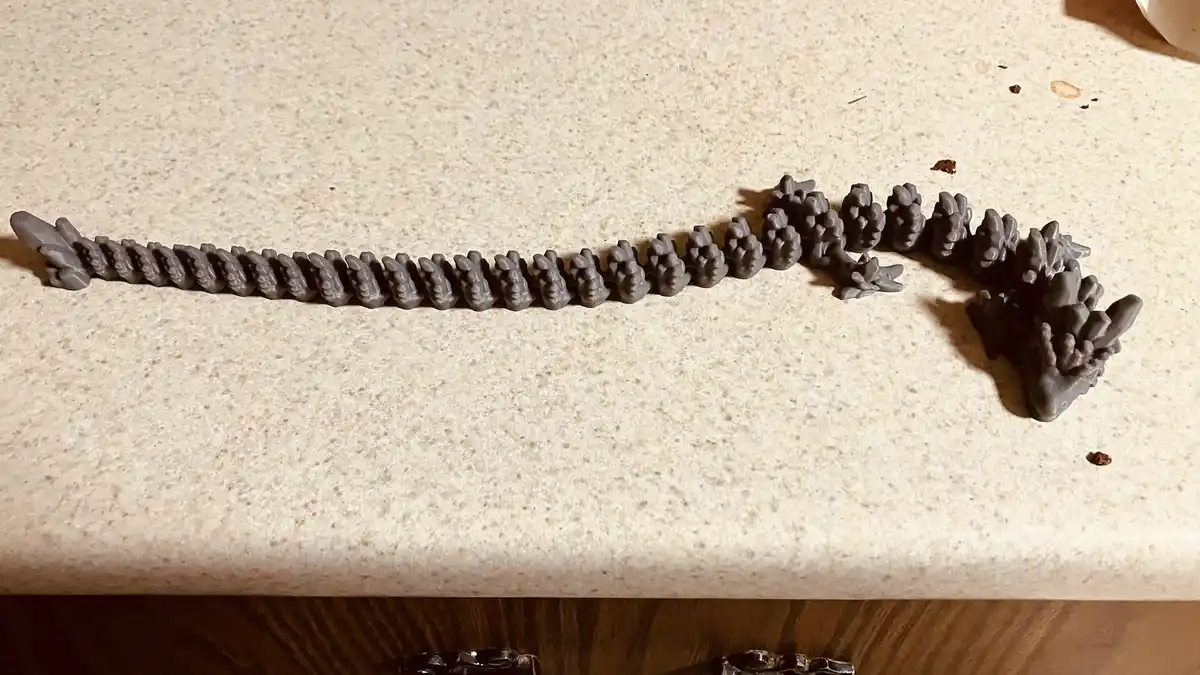 A gray 3D-printed dragon skeleton model lies on a speckled beige countertop, its spine and head clearly visible. The…
