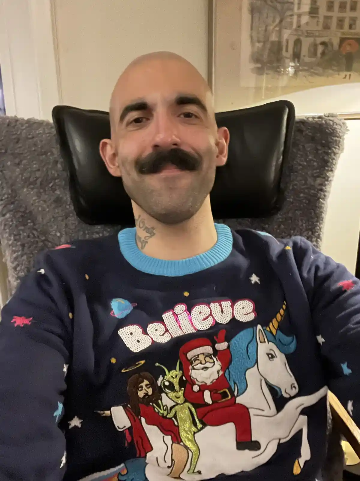 A bald man with a thick mustache sits in a chair wearing a dark blue, sparkly Christmas sweater with a chaotic design…