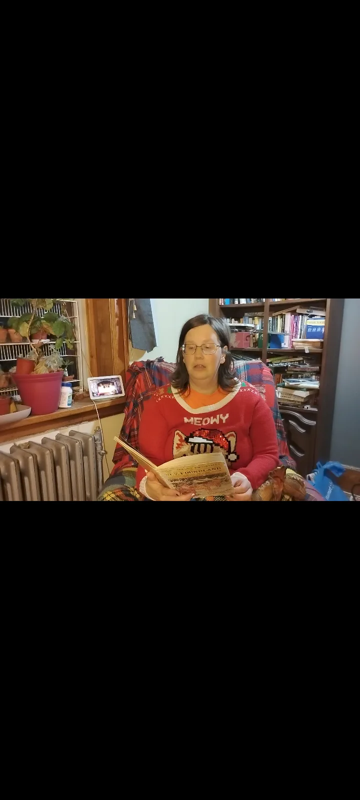 A woman wearing a red 'MEOWY' sweater sits in a chair, holding an open book titled 'MY FORDLAND'. She has glasses and a…