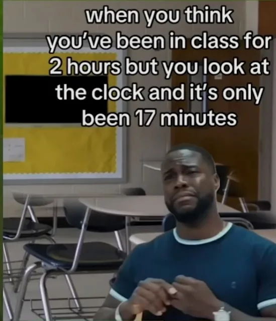 A man with a weary, disappointed expression sits in a classroom with empty desks and chairs around him. Behind him is a…