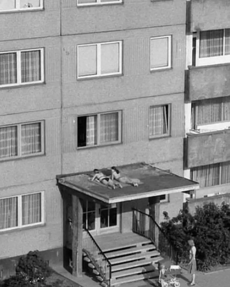 A black-and-white photo shows two people lying on a flat roof of a multi-story apartment building, appearing to sunbathe…