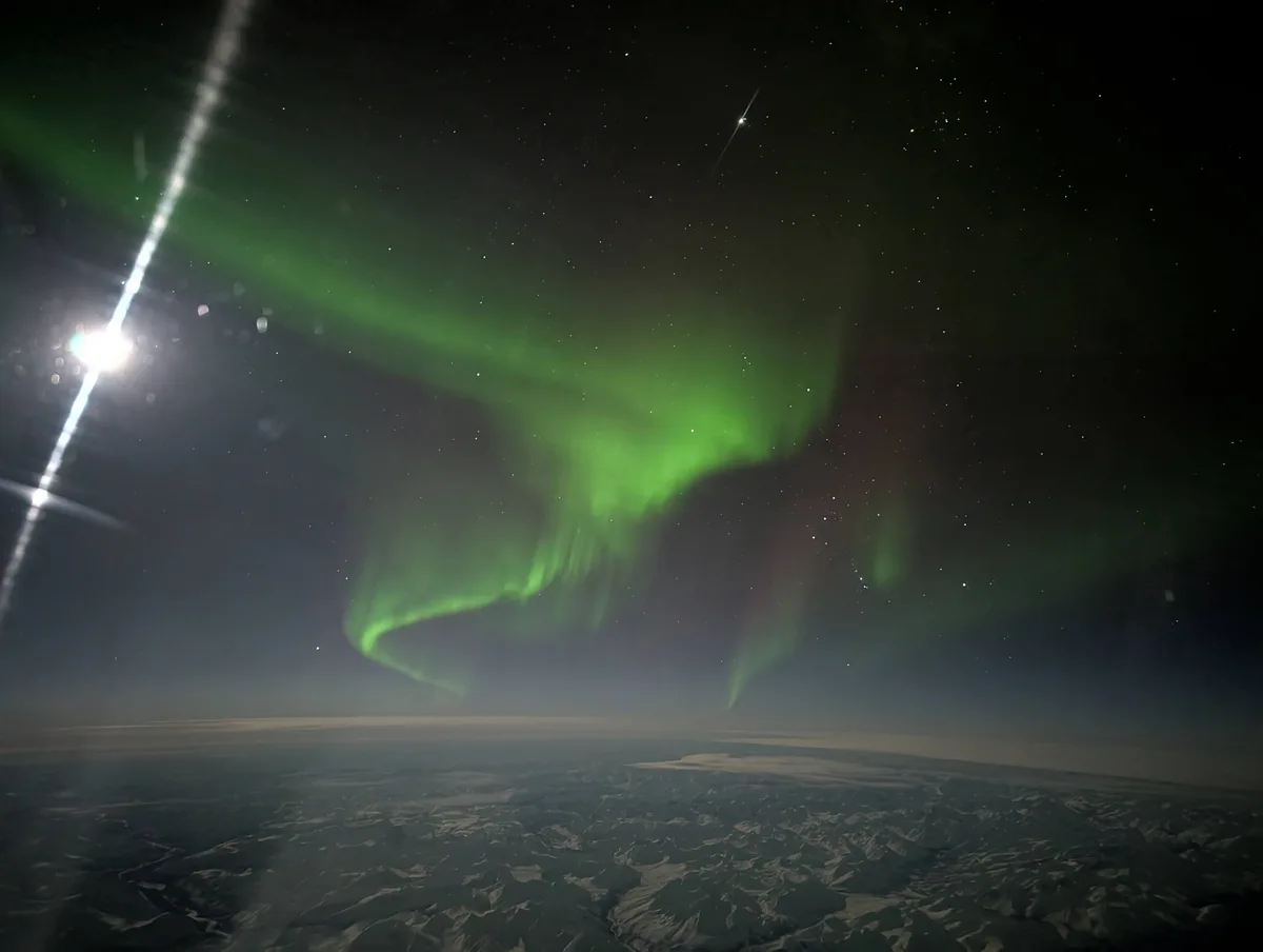 A photograph taken from an airplane window shows a vibrant green aurora borealis dancing across a starry night sky above a…