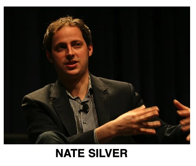 A man with short brown hair wearing a dark pinstripe suit jacket over a checkered shirt is captured mid-gesture, seemingly…