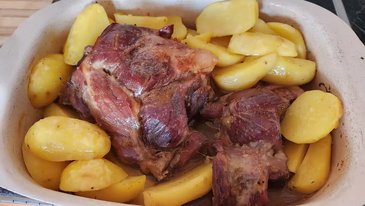 A close-up photo shows a white ceramic baking dish filled with cooked roast beef and golden-yellow potato wedges in a…