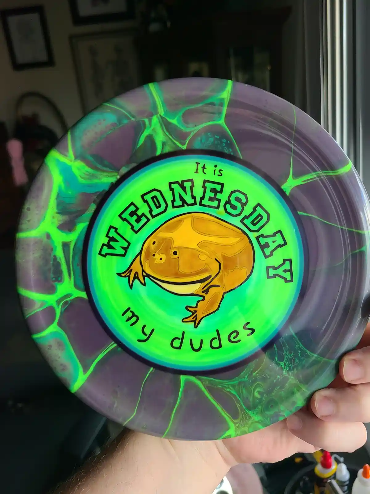 A hand holds a custom disc golf disc with a marbled purple and neon green design. The center features a cartoon frog and the…