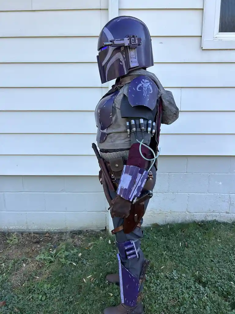 A child dressed in a detailed, weathered Mandalorian armor costume, standing in profile against a white house wall. The…