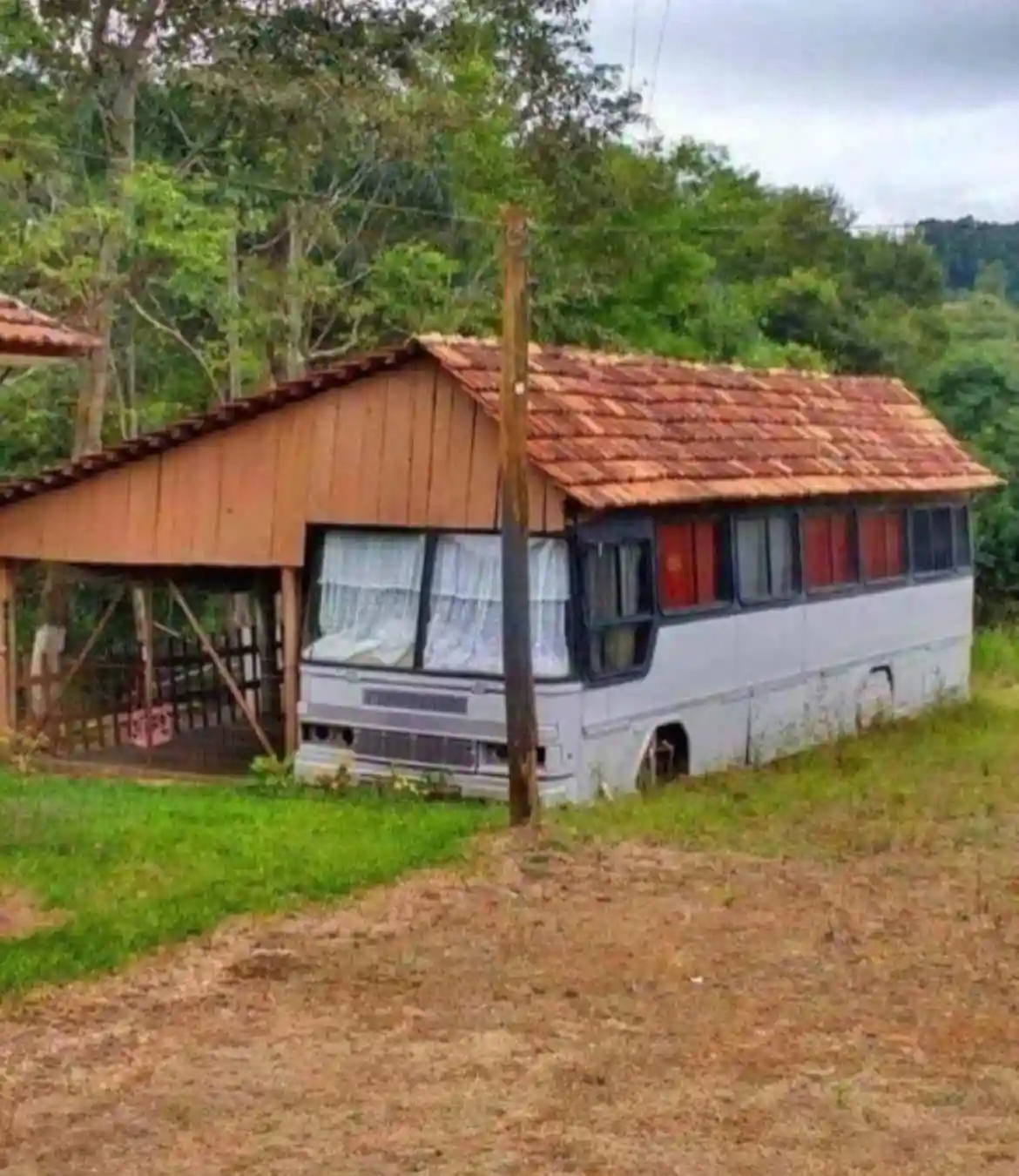 A white bus has been modified into a house, with a wooden roof and porch added on top, set in a grassy, wooded area. The…