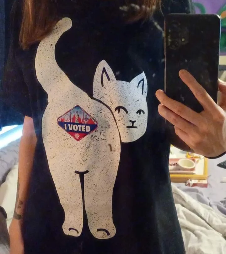 A person takes a mirror selfie wearing a black t-shirt with a large white cartoon cat graphic. The cat's body has a red…