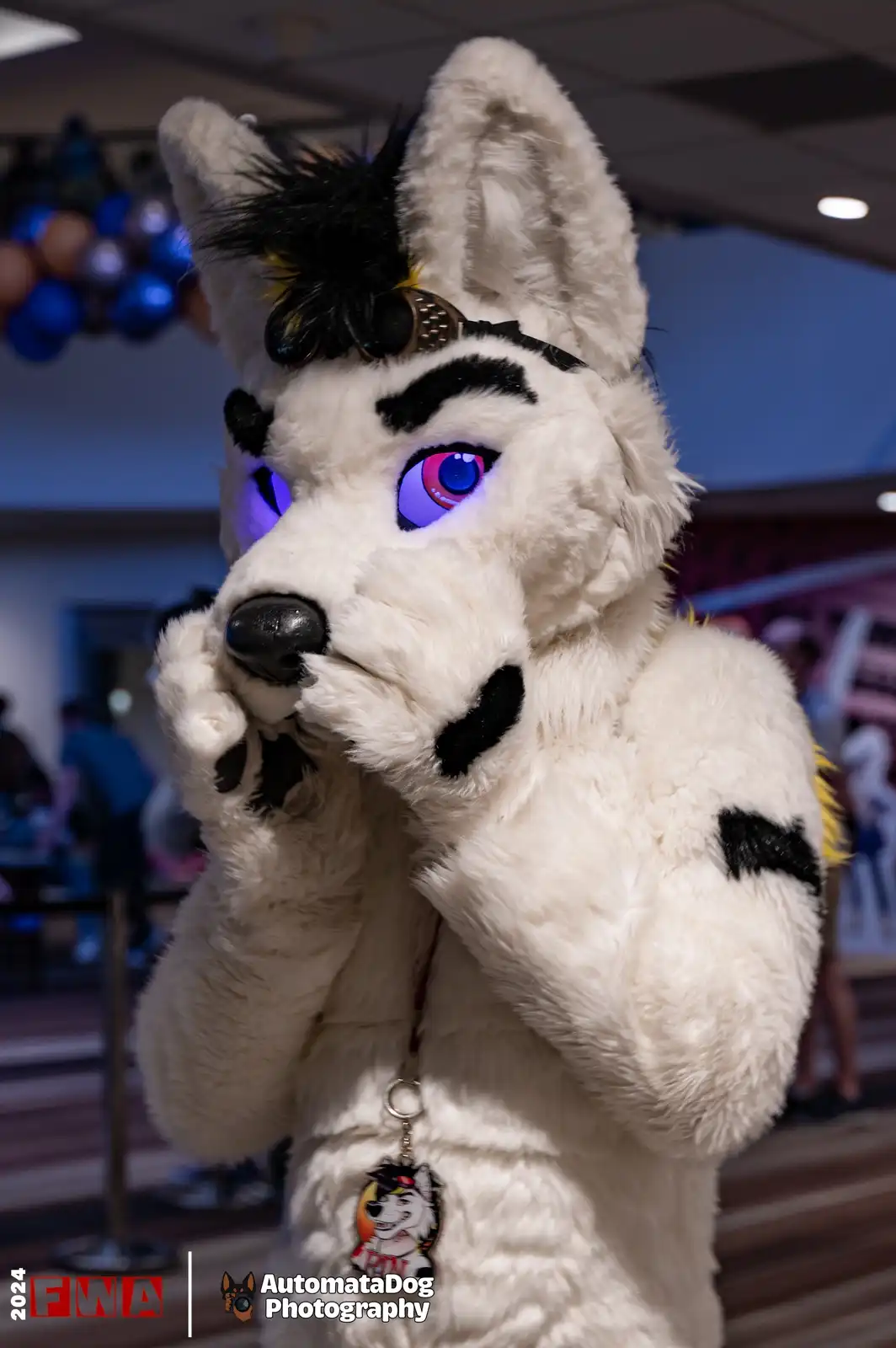 A person in a detailed white fursuit with black markings, glowing purple eyes, and a black mohawk, holds their paws near…
