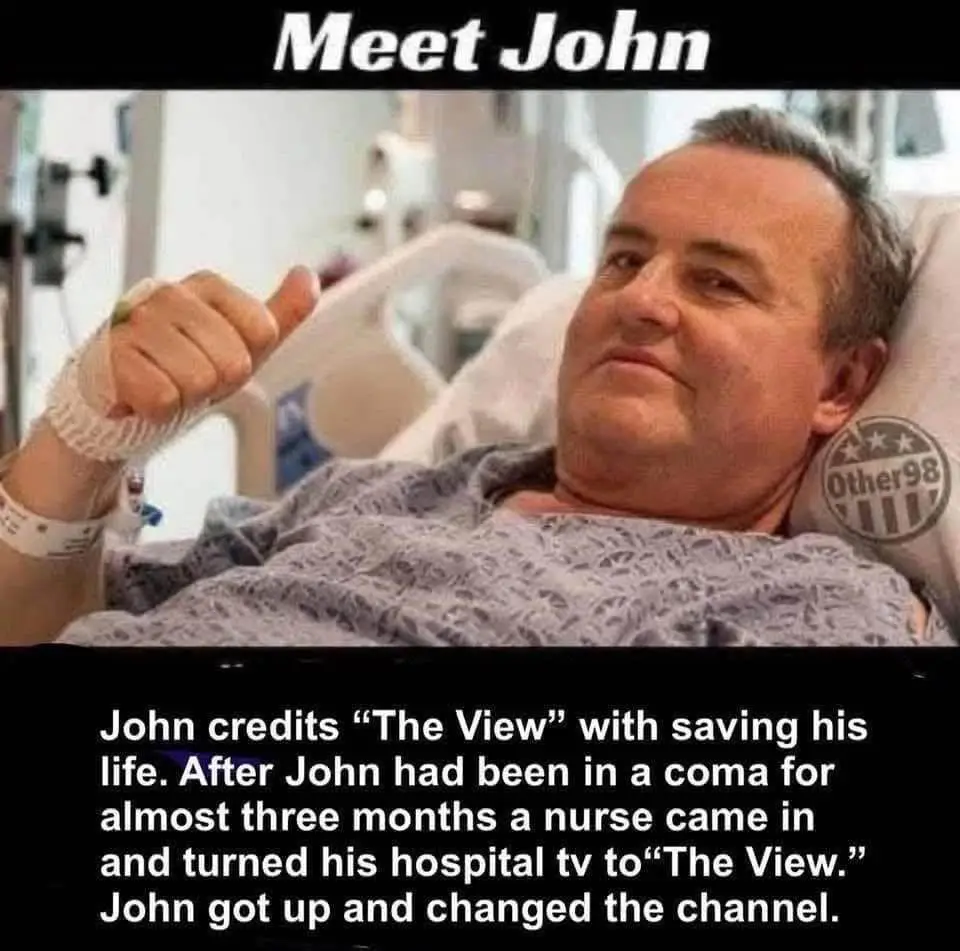 A man named John lies in a hospital bed with a bandaged hand, giving a thumbs-up while looking at the camera. The image has…
