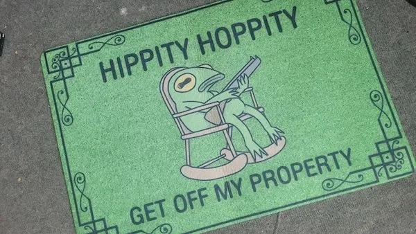 A green doormat with ornate borders features a cartoon frog lounging in a rocking chair while holding a shotgun. The text…